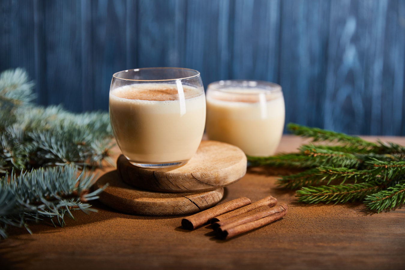 tasty eggnog cocktail on round wooden boards near spruce branches and cinnamon sticks on blue textured background