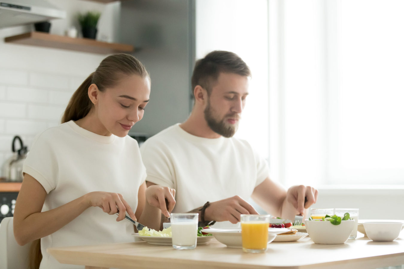 Young happy couple eating with knife and fork together at home, man and woman having dinner at kitchen dining table, tasting healthy balanced delicious yummy breakfast, enjoying organic meal